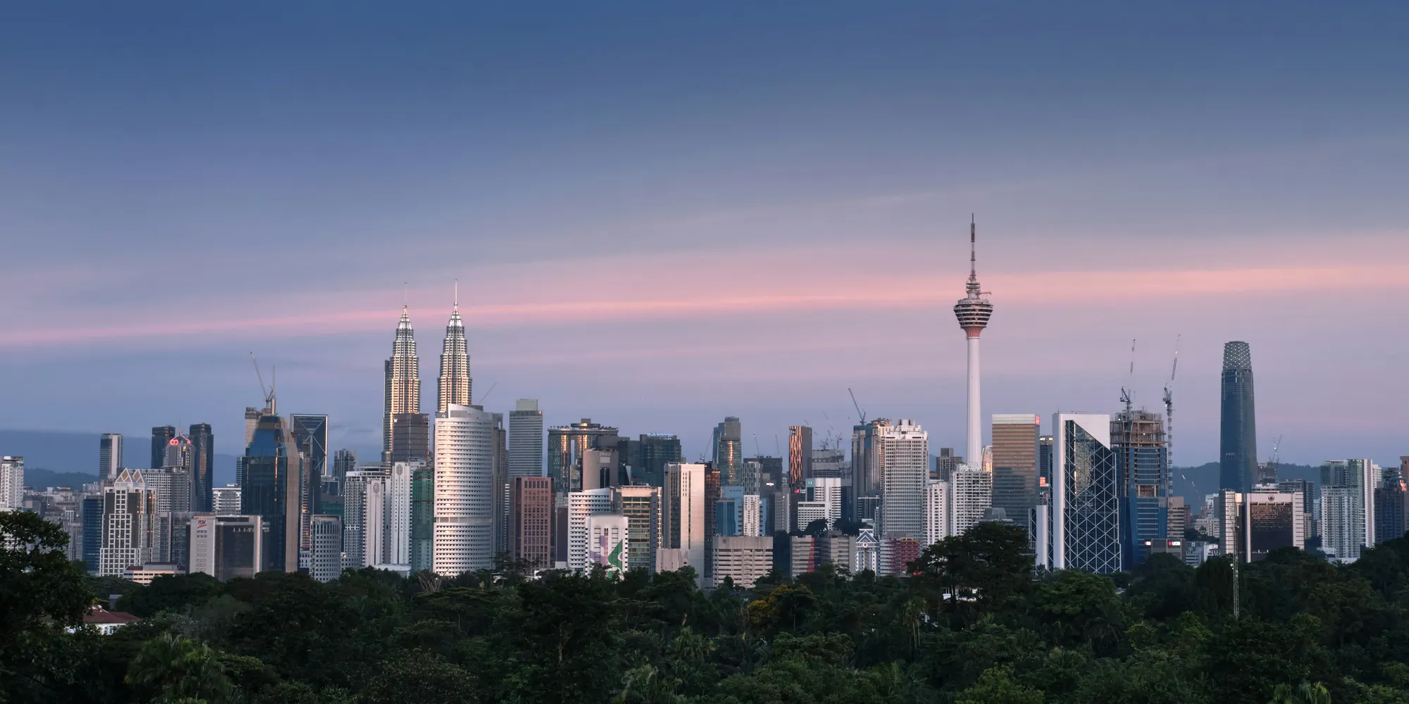 The Kuala Lumpur skyline at dusk, with the Petronas Towers and KL Tower above a band of rainforest